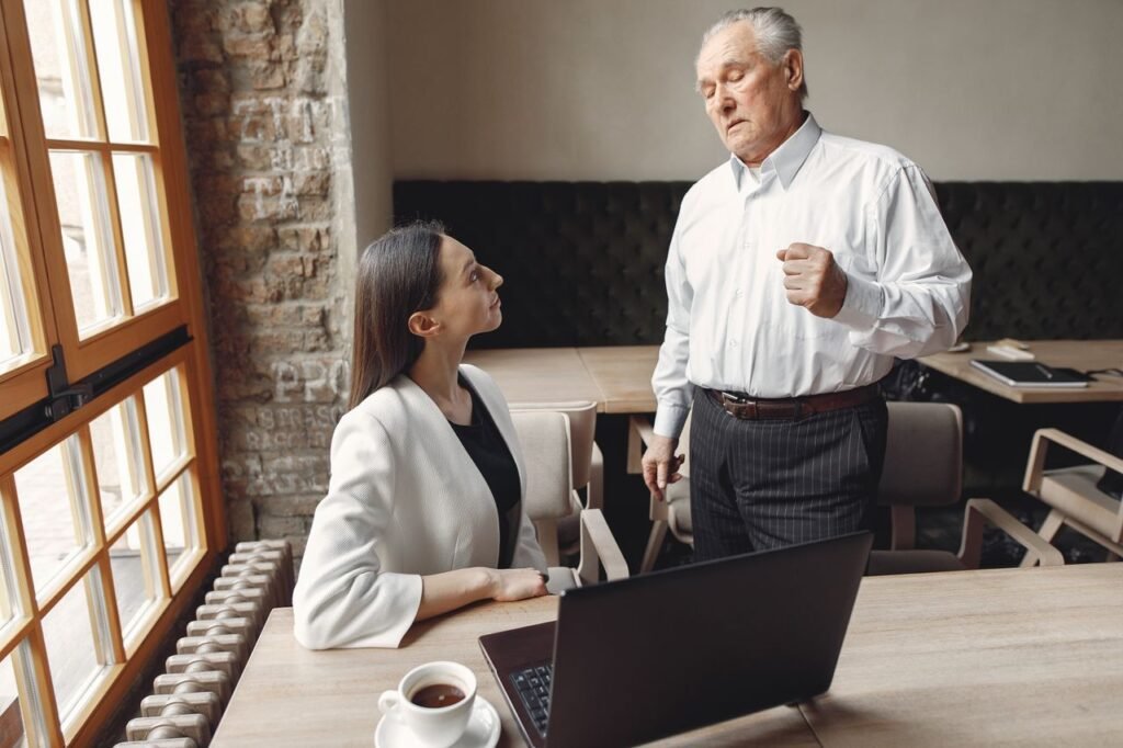Aged businessman in formal wear talking with well dressed female employee with laptop and cup of coffee about business issue in modern office